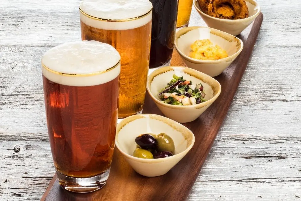 A selection of four beers with small bowls of snacks on the side that all sit stop a wooden table.