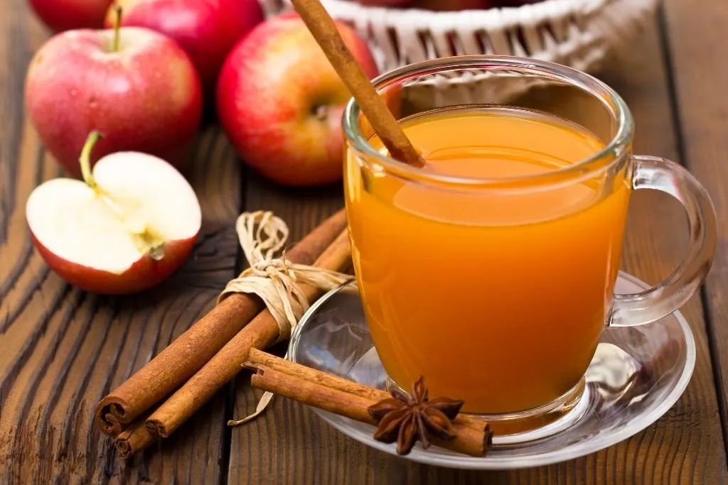 A glass of hot apple cider on a picnic table with cinnamon and freshly cut apples around it. 