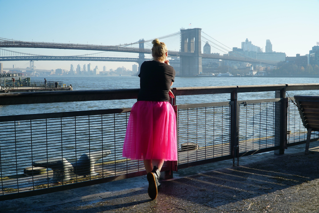 Me standing in a pink skirt looking out at the Brooklyn Bridge from South Street Seaport during January in NYC. 