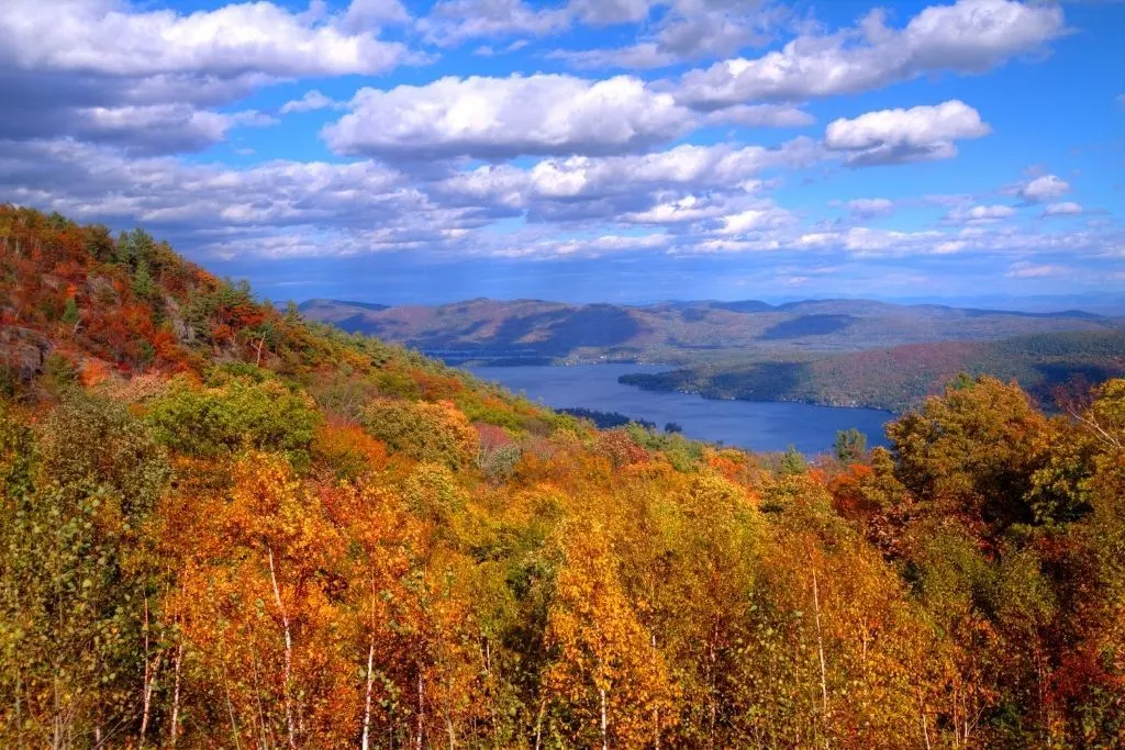 Aerial view of Lake George from the top of a mountain in the fall with vibrant foliage in the foreground. Catching the fall colors is one of the popular things to do in Lake George.