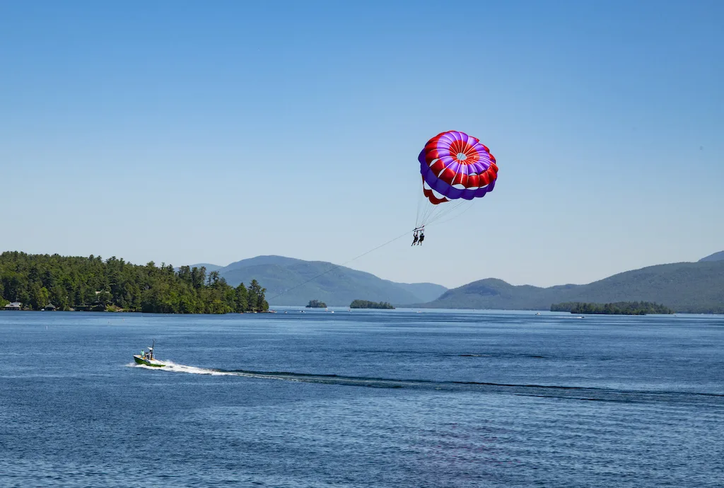 View of Lake George and mountains with two parasailing persons in the foreground. Parasailing is one of the adventurous things to do in Lake George.