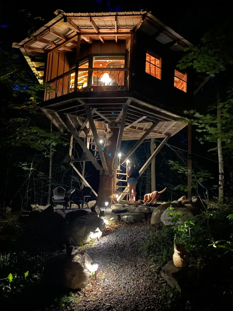 Woman and dog headed up the spiral staircase to this octagon-shaped treehouse which is all lit up in the evening. 
