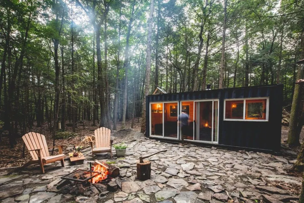A black container cabin that is now a tiny house New York getaway with an outdoor fire pit and two Adirondack chairs. 