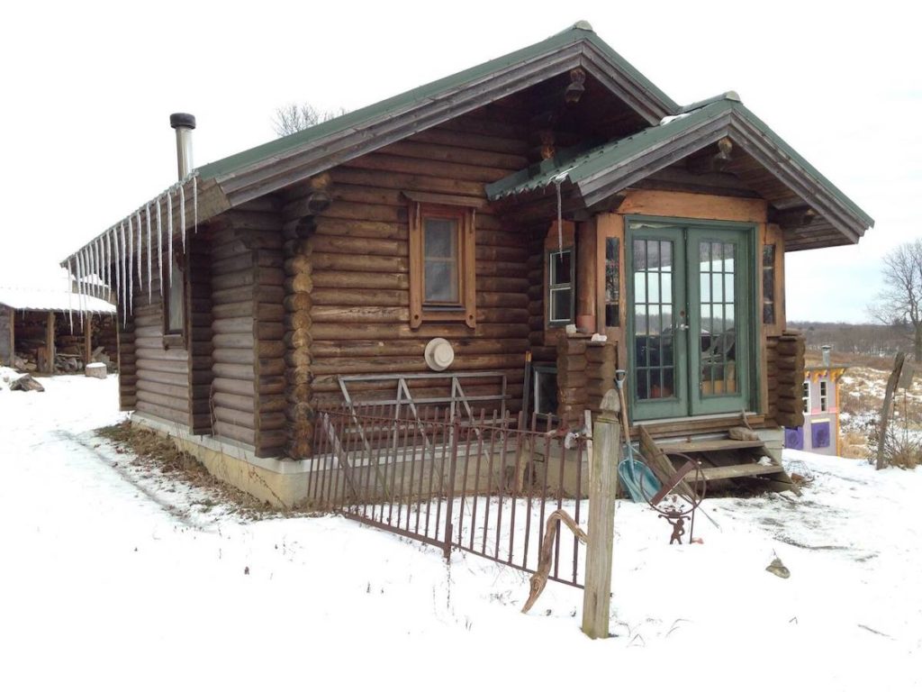 Tiny house in "Karenville" near Ithaca Ney York. It's covered in snow and is a log cabin with green doors. 