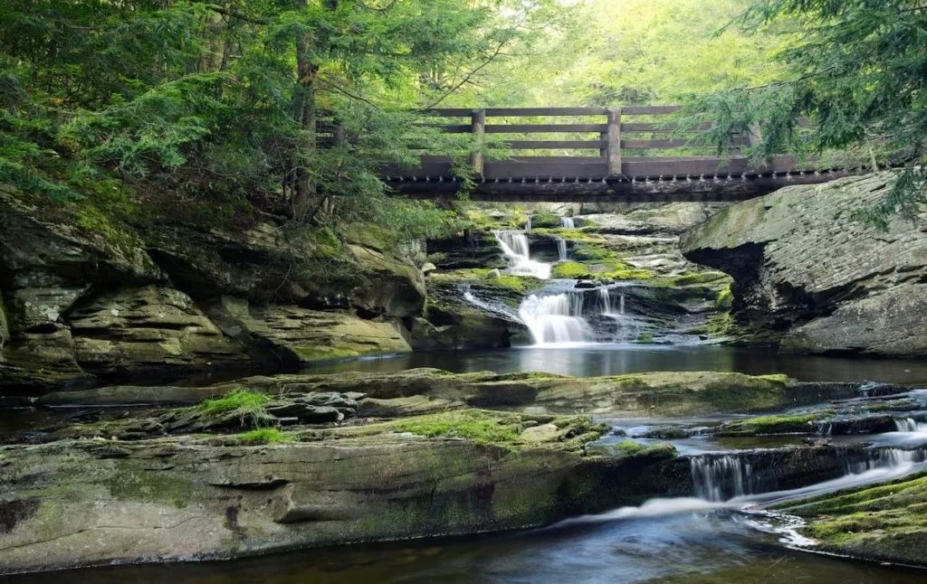 Vernooy Falls with a wooden bridge and mossy stone banks in the Catskills is one of the fun swimming holes in NY