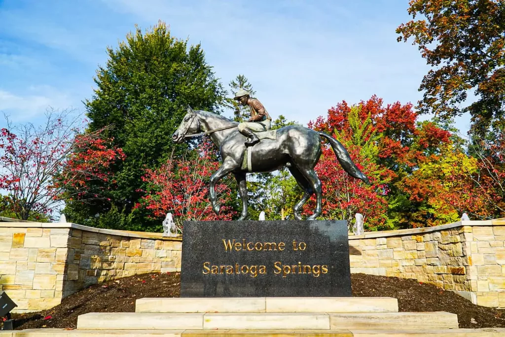 Welcome to Saratoga Springs sign with a jockey and horse on top at Congress Park. and the colorful red leaved trees in the background is one of the popular things to do in Sraatoga springs