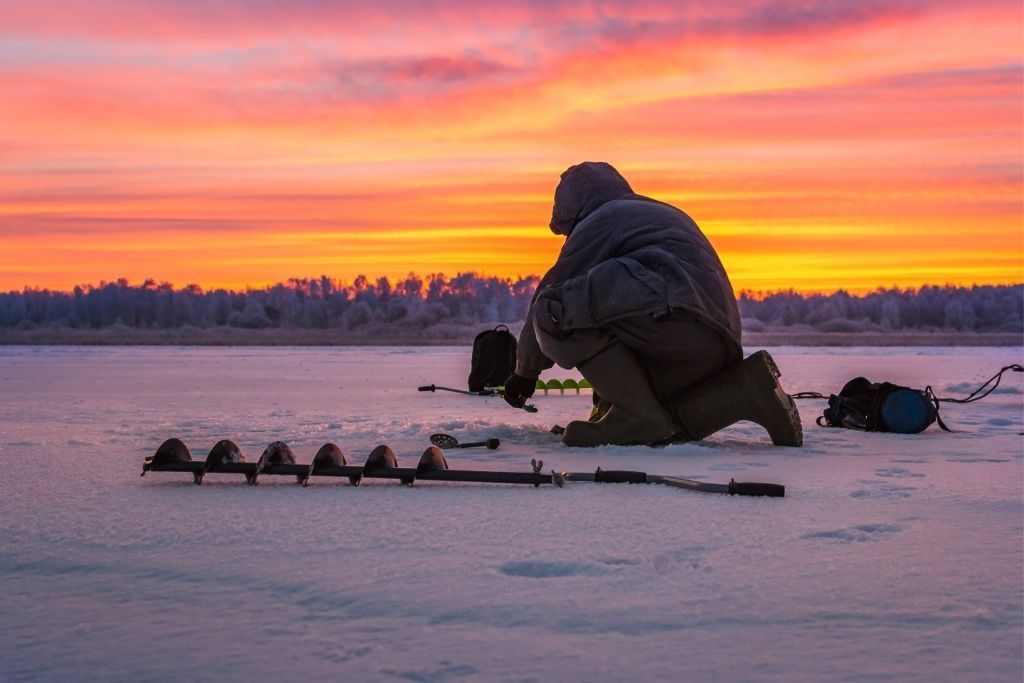 Man going ice fishing on a lake in the winter as the sun sets. One of the best things to do in winter in Lake George/ 