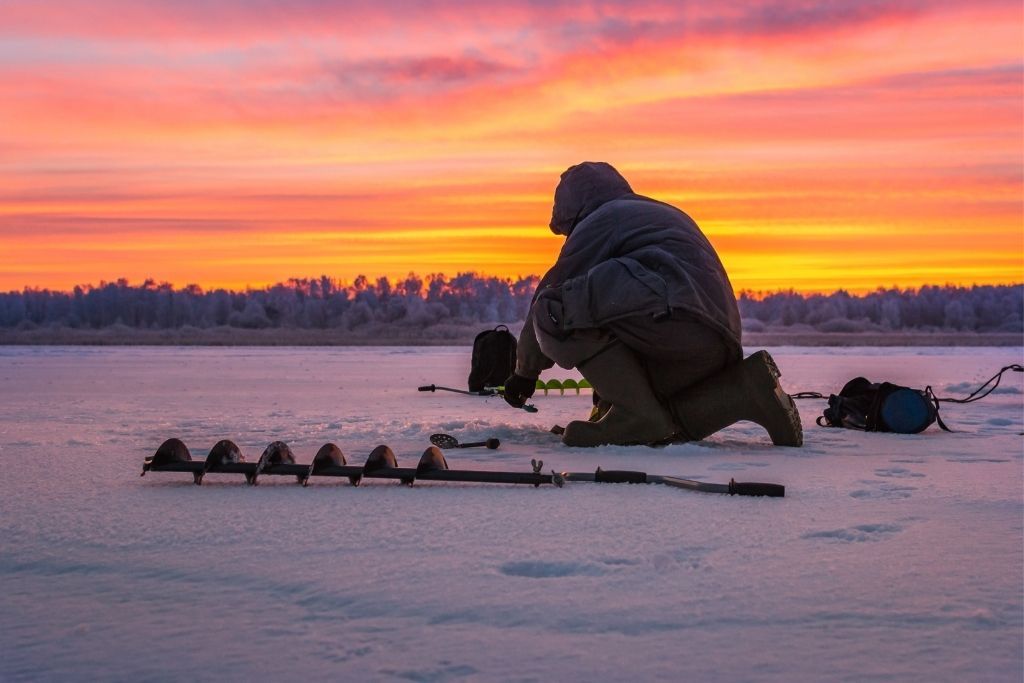 Man going ice fishing on a lake in the winter as the sun sets. One of the best things to do in winter in Lake George/