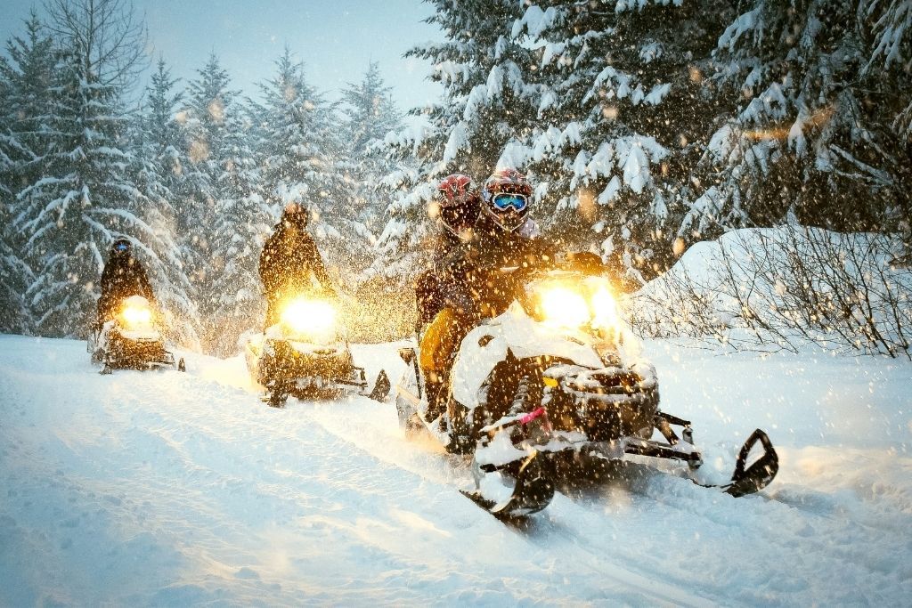 A group of people going down a snowy trail in the evening while riding snow mobiles. Easily one of the best things to do in winter in Lake George.