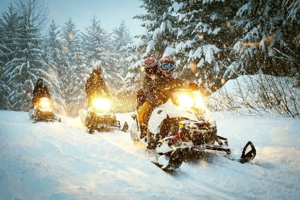 A group of people going down a snowy trail in the evening while riding snow mobiles. Easily one of the best things to do in winter in Lake George. 
