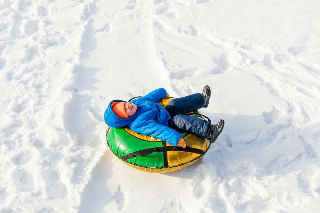 A small boy in a blue snow suit is riding on a green and yellow inner tube and along the snow. 