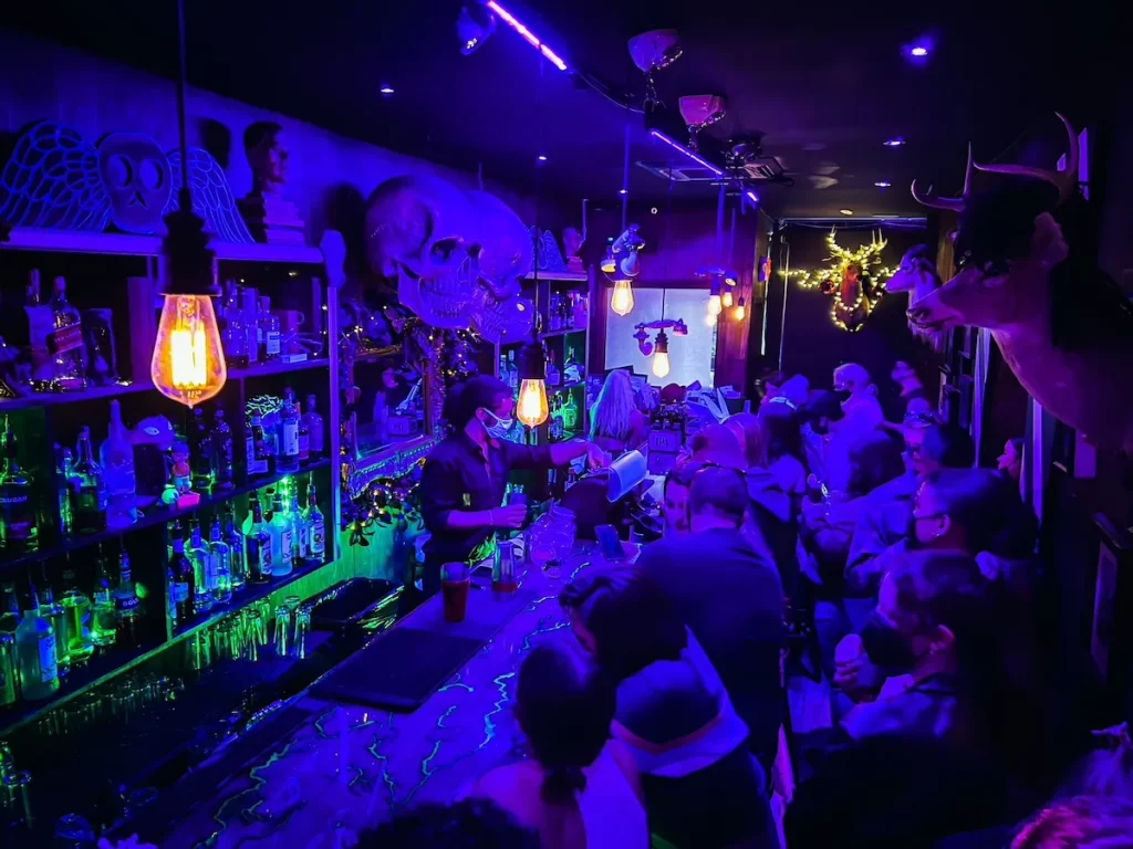 View of the black light and bar that can be found inside Beetle House. People stand around the bar and their are skulls on the walls. 