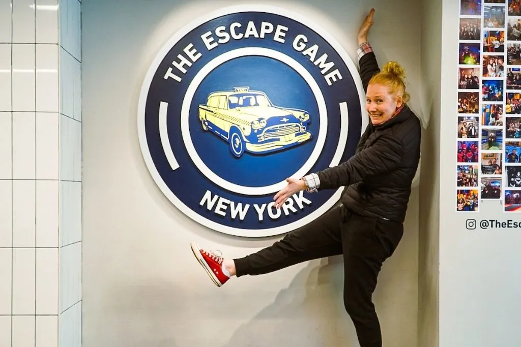 Me standing in front of a sign for the dark blue escape game logo with a car symbol in it, one of the best things to do in NYC for your birthday. 