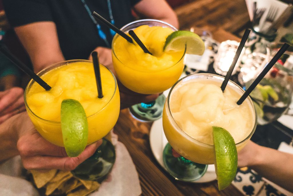 A view of three margaritas. One is lighter and two are frozen mango margaritas. All come with black straws and lime wedges. 