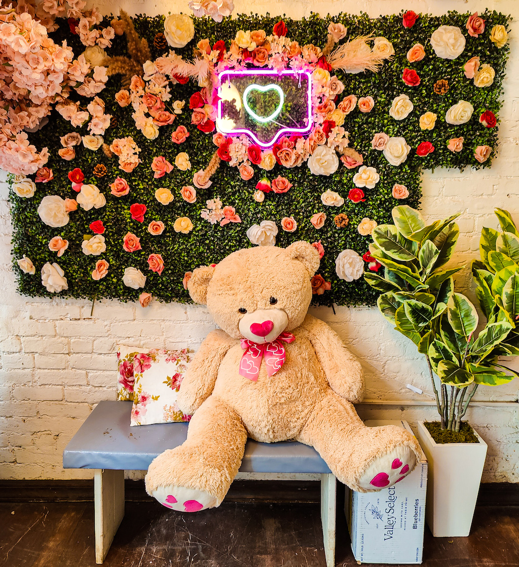 A giant teddy bear sitting in front of pastel flowers on a bench inside eat pretty cafe. 
