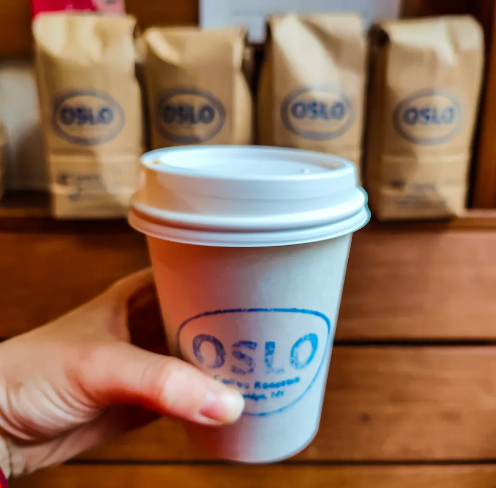 Holding a to-go cup at Oslo Offee Roasters in front of some bags of ground coffee. 