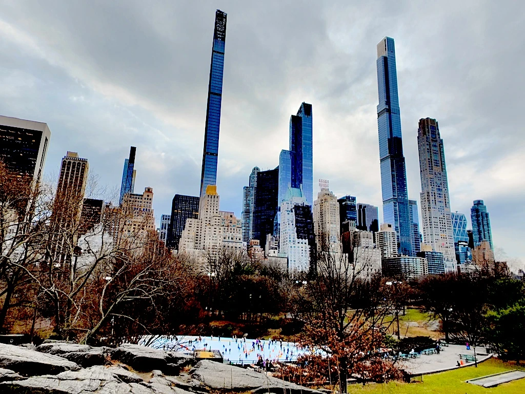 A view of central park in winter with skyscrapers in the background and Wollman Rink in the middle with ice skaters skating in the winter and a cloudy sky. 