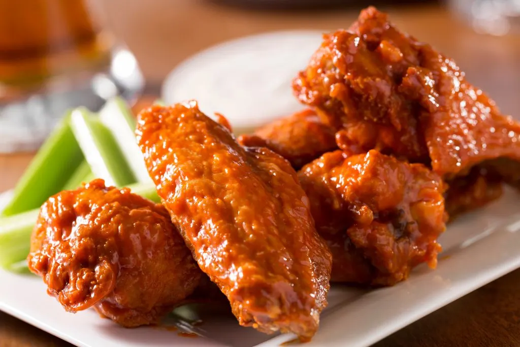Five delicious chicken wings smothered in red sauce that are sitting on a while plate with a side of ranch in the back and some celery from one of the best Seneca Lake Breweries. 