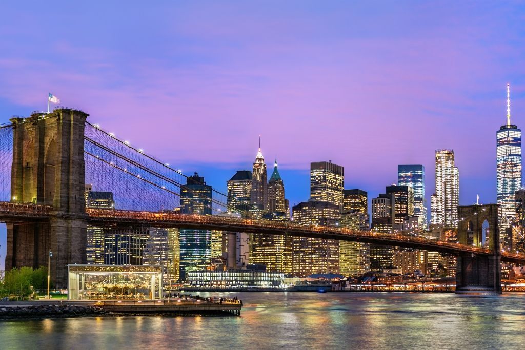 View of the Manhattan skyline at twilight. The sky is blue and purple and this shot was taken from Brooklyn Bridge Park. You can see the Brooklyn Bridges and famous Manhattan skyscrapers l it up in the background during one of the best NYC night tours. 