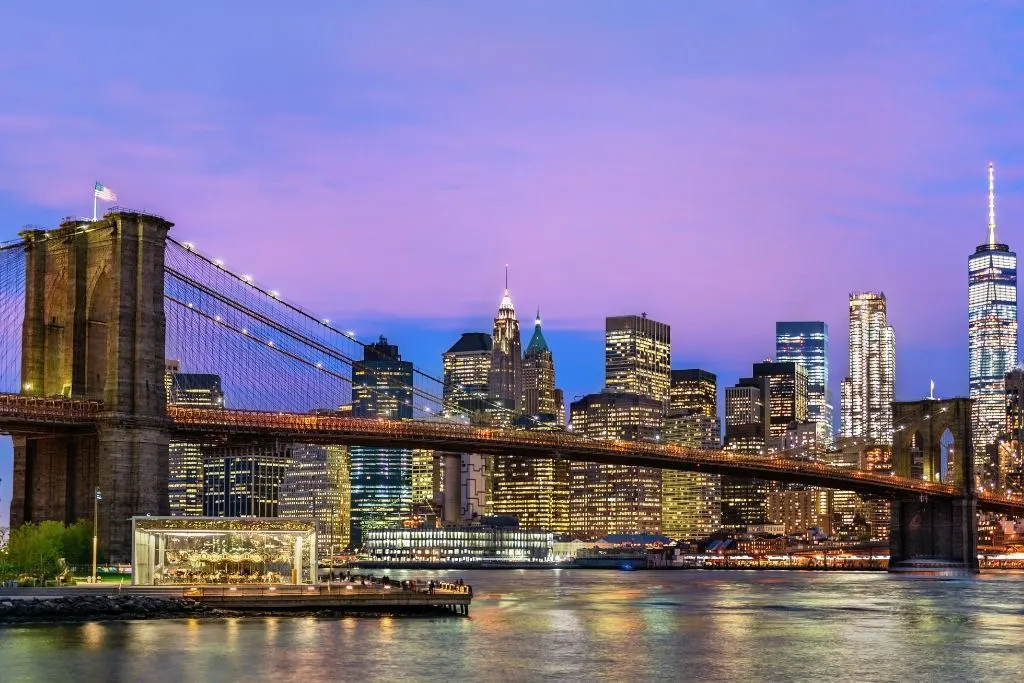 View of the Manhattan skyline at twilight. The sky is blue and purple and this shot was taken from Brooklyn Bridge Park. You can see the Brooklyn Bridges and famous Manhattan skyscrapers l it up in the background during one of the best NYC night tours. 