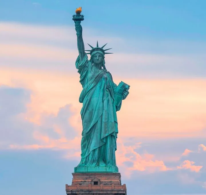 Up close view of the Statue of Liberty in green while she holds her torch with a yellow flame. The background is at twilight.