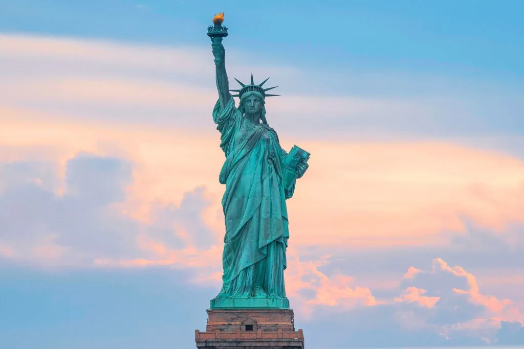 Up close view of the Statue of Liberty in green while she holds her torch with a yellow flame. The background is at twilight. 