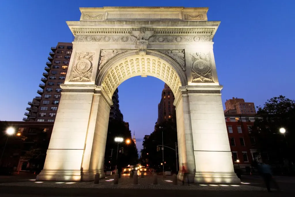 Giant white marble arch in Washington Park all lit up in the evening. Through the arch you can see down fifth avenue and see taxis on the road at night. 