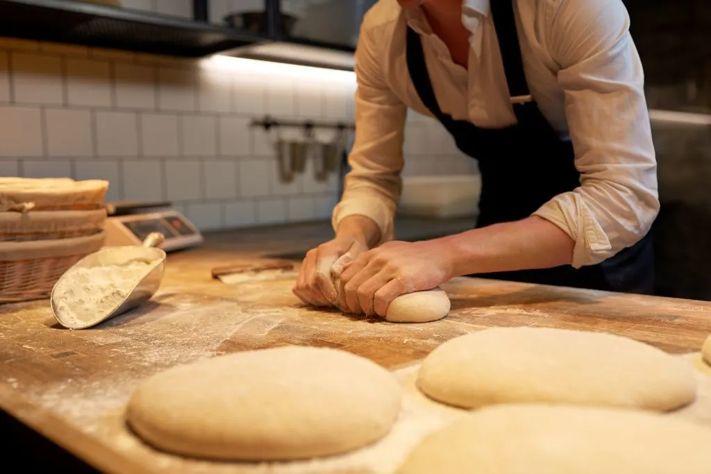 Baker in a white shirt and dark apron has both hands on dough and is shaping it into fresh bread that you can enjoy during your Beacon itinerary,