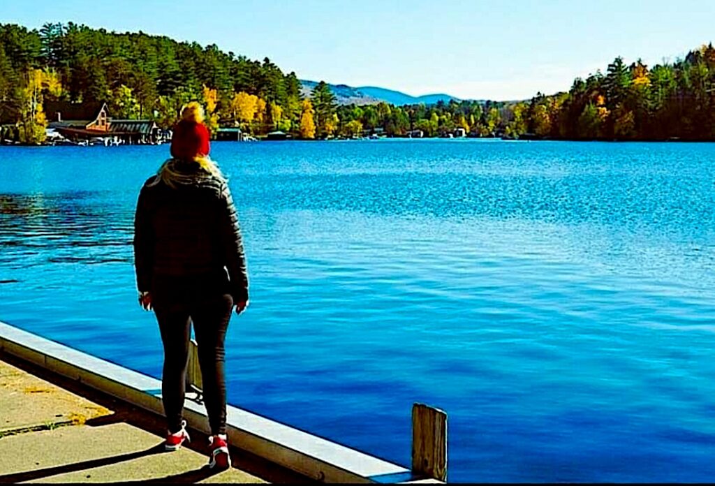 Standing in front of Saranac Lake in the fall with a red hat and a black jacket. Just looking out at the lake with fall foliage in the background. 