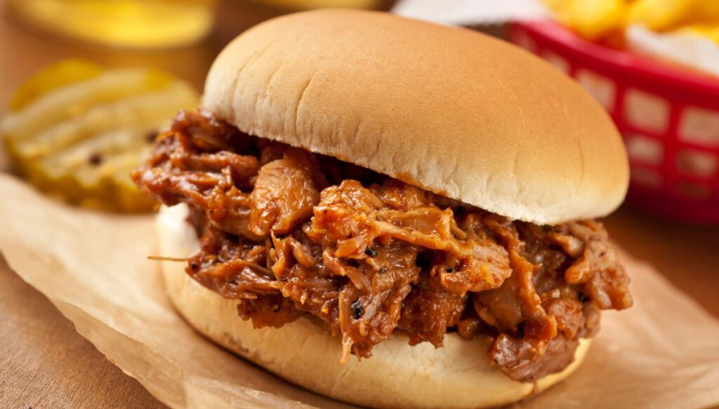 A bbq pulled pork sandwich on a bun with pickles and fries in the background.