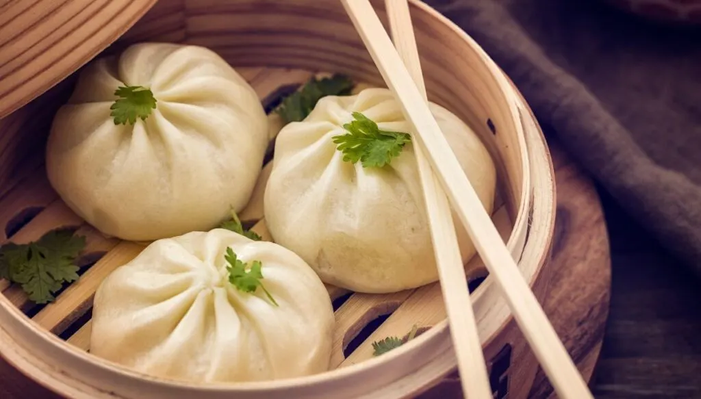 A view of three dim sum dumplings in a wooden steaming basket with chopstikcs on top. 