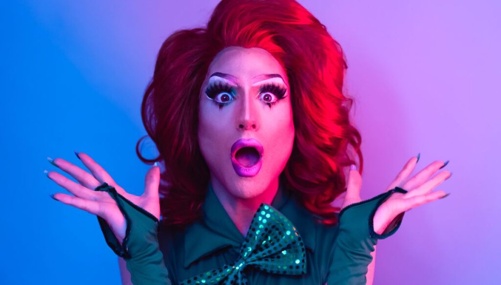 A drag queen with red hair and a green shirt with a bow tie with a pink/purple background at the best drag brunch NYC has to offer. 