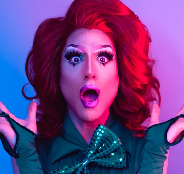 A drag queen with red hair and a green shirt with a bow tie with a pink/purple background at the best drag brunch NYC has to offer.