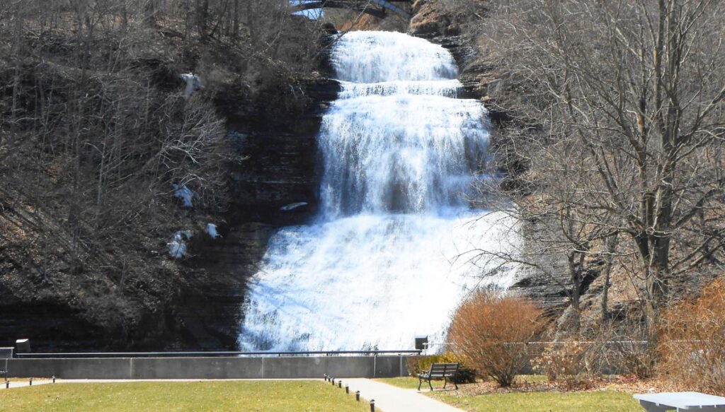 View of the majestic Montour Falls in the center of the picture. It is winter and surrounded by bare trees and has a fence in front with a grassy area and a path that leads to the falls and one of the best things to do in Watkins Glen, NY. 