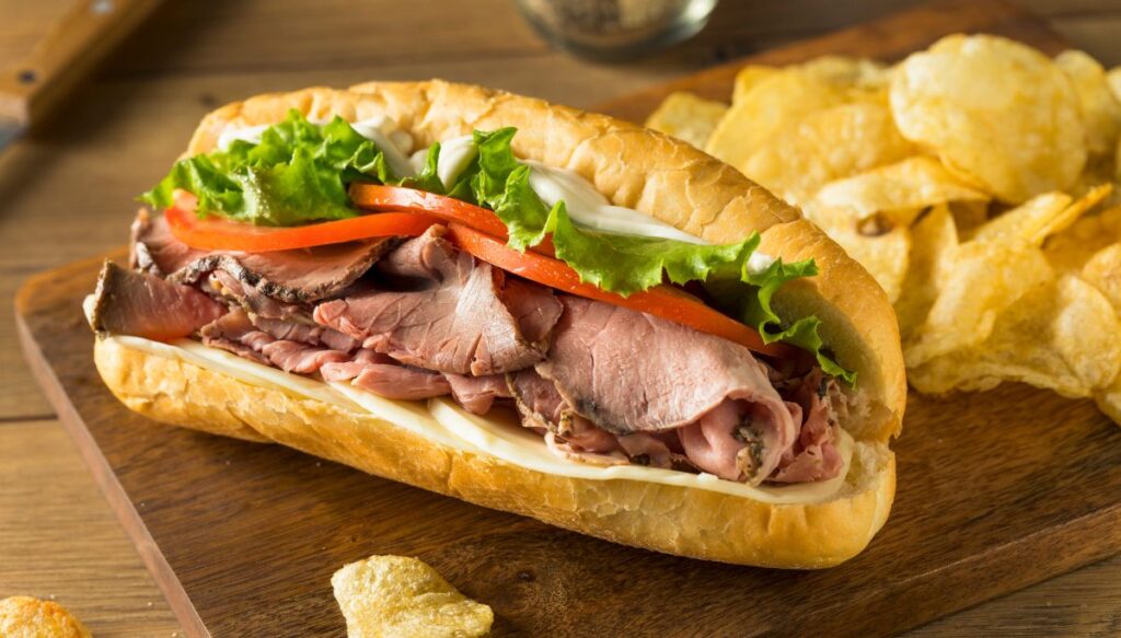 A roast beef sandwich on a sub roll with lettuce, tomoto, and chips in the background. 