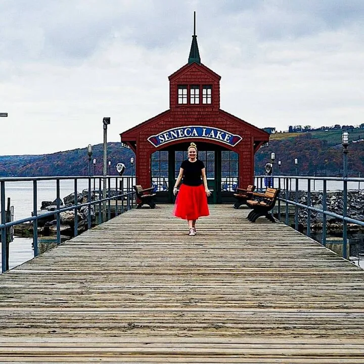 A picture of me in a black t-shirt and red skirt walking towards the camera along a wooden boardwalk with a red steepled building in the background, on the lake, that has a blue sign that says Seneca Lake. This is a great spot to enjoy some fo the best things to do in Watkins Glen.
