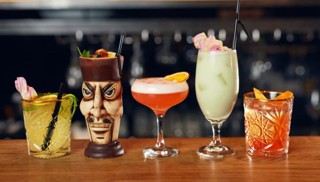 A view of five different tropical themed drinks on a wooden bar with the full bar in the background. 