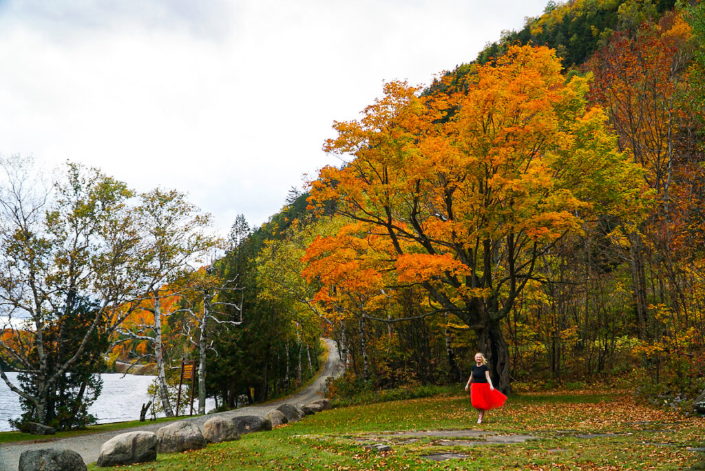 Me exploring Lake Placid. There is a tree with orange leaves in the background as I walk on grass in a black t-shirt and red skirt. There is a dirt road to the left and small lake just behind it as I explore the area during the best time to visit Lake Placid. 