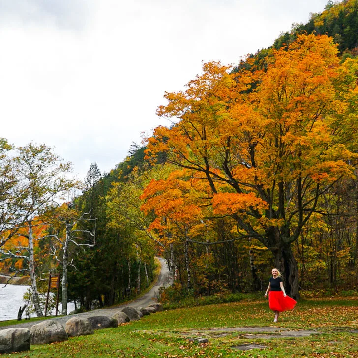Me exploring Lake Placid. There is a tree with orange leaves in the background as I walk on grass in a black t-shirt and red skirt. There is a dirt road to the left and small lake just behind it as I explore the area during the best time to visit Lake Placid.