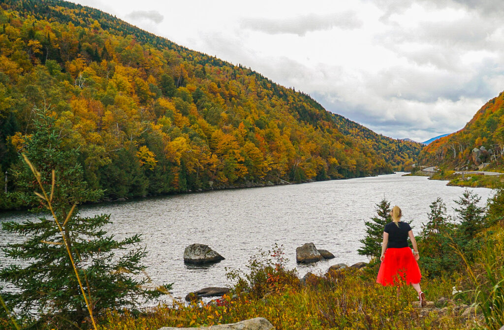 Me with my back to the camera in a red skirt and black t-shirt. You can see the leaves of the evergreen trees changing around a lake near Lake Placid. The is one of the best months to visit Lake Placid. 