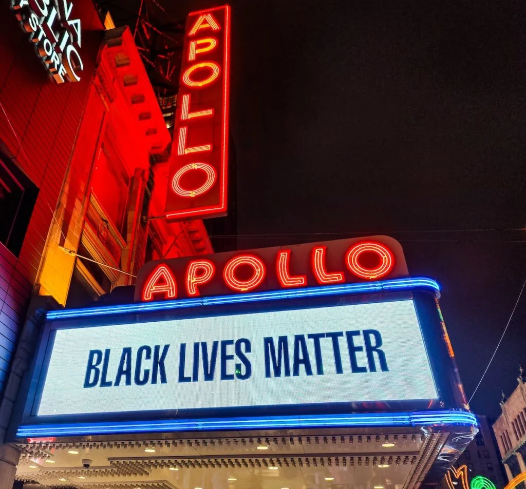 Vertical and horizontal red letters say. "Apollo" above the marquis at the Apollo Theater in Harlem. The white sign says "black lives matter". 