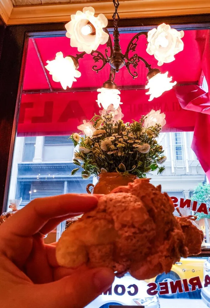 Me holding a croissant at Balthazar bakery. You can see vintage lights hanging from the ceiling and a red awning through the window in the background. There is also a green plant with white flowers right behind me. 