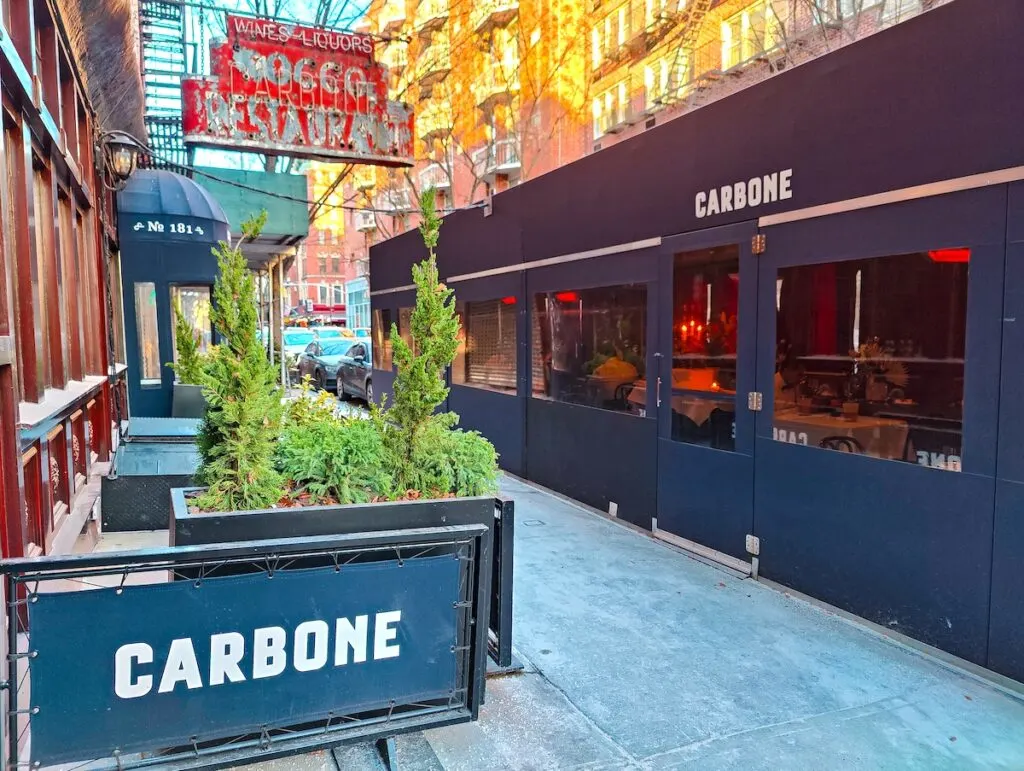 The exterior of Carbone. You can see a red sign above green potted plants and blue signs on the ground. There is also a square tent with tables and 