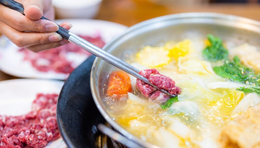 A person is holding metal chopsticks and using them to cook meat in a hot pot with soup and veggies. 