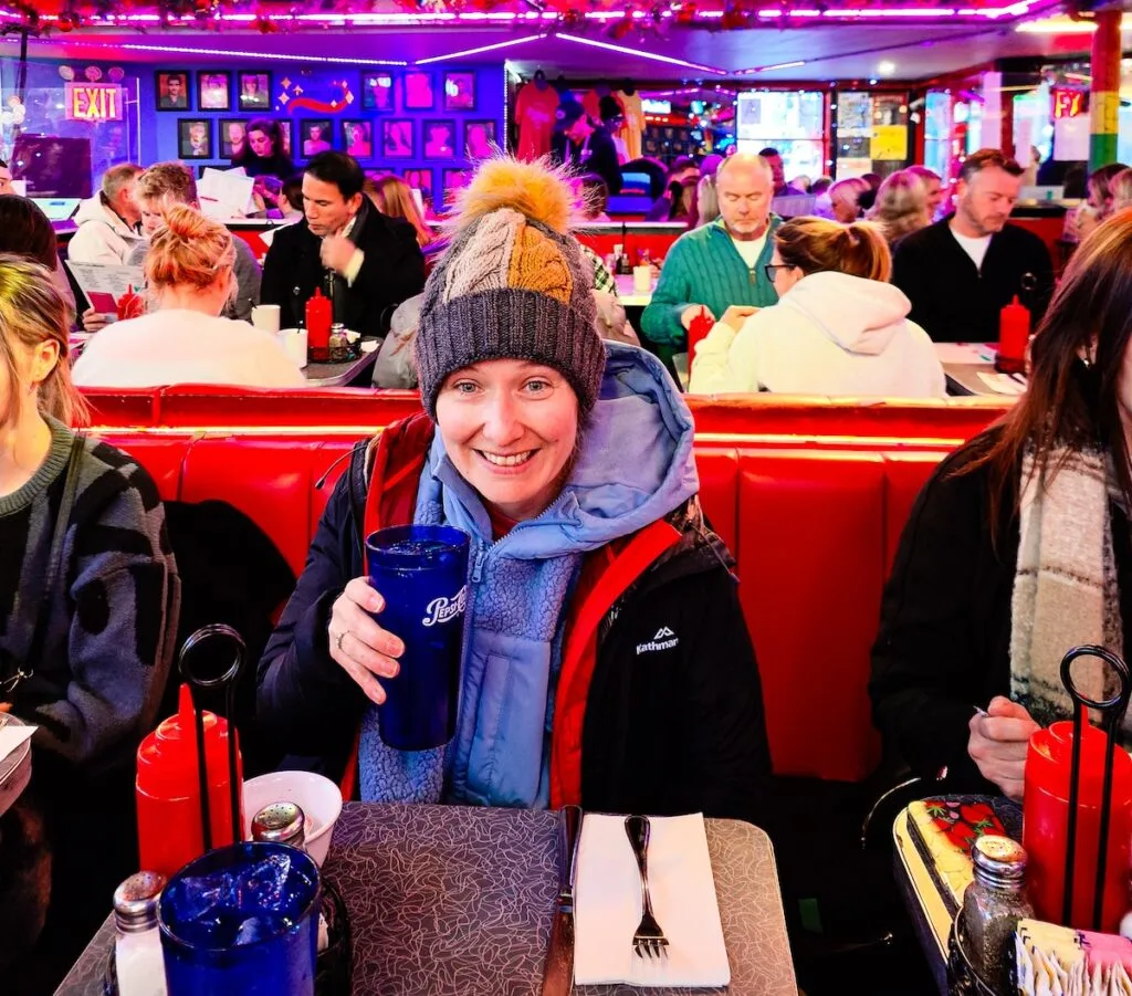Me sitting in a jacket and hat while sitting at a red booth inside Ellen's Stardust Diner. I am holding a blue glass with 