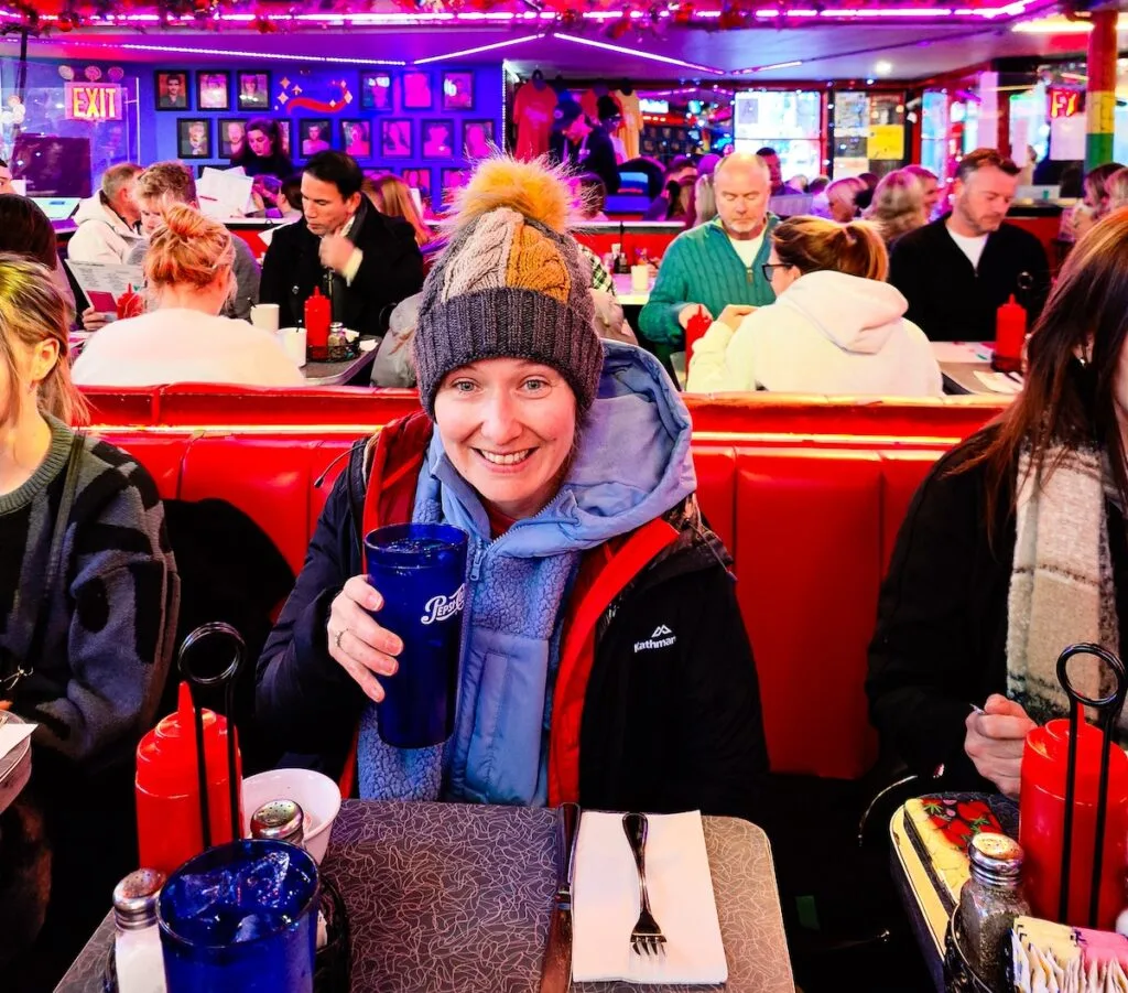 Me sitting in a jacket and hat while sitting at a red booth inside Ellen's Stardust Diner. I am holding a blue glass with "pepsi" on it and am enjoying one of the fun restaurants in NYC. 