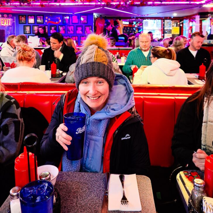 Me sitting in a jacket and hat while sitting at a red booth inside Ellen's Stardust Diner. I am holding a blue glass with 