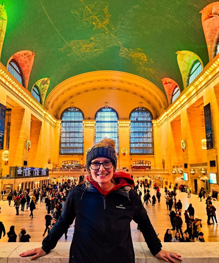 A picture of me standing on the platform in front of the ceiling in Grand Central with a black coat, a hat, and my hands on the railing. 