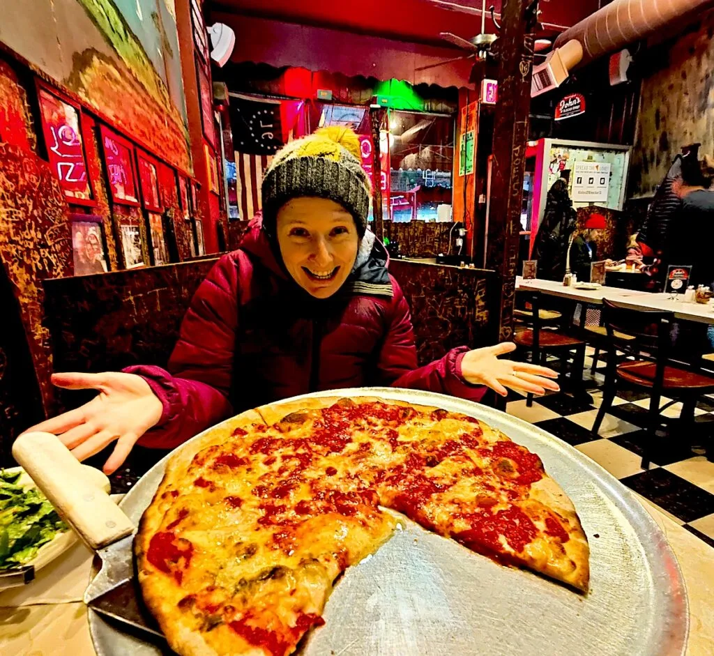 Me sitting behind an original pizza pie on a metal tray with a pizza server on the left. I have my hands out behind the pizza and am wearing a pink coat and yellow hat at John's of Bleecker Street.