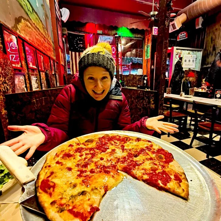 Me sitting behind an original pizza pie on a metal tray with a pizza server on the left. I have my hands out behind the pizza and am wearing a pink coat and yellow hat at John's of Bleecker Street.